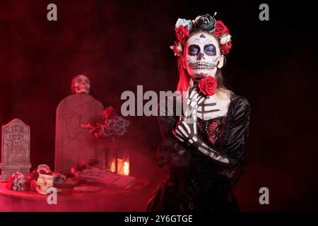 Frau posiert mit roter Rose in der Friedhofsszene, während sie Skelett-Make-up trägt, um dios de los muertos zu feiern. Makabre Dame, gekleidet als La Catrina Charakter am Tag des Todes im nebeligen Studio Stockfoto