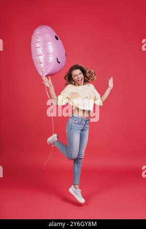 So glücklich! Volle Länge einer attraktiven jungen lächelnden Frau, die einen herzförmigen Ballon hält und schreien, während sie vor rosa Hintergrund springt Stockfoto