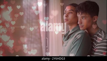 Bild von glitzernden Herzen über glücklichen, vielseitigen, schwulen männlichen Paaren, die aus dem Fenster schauen und umarmen Stockfoto