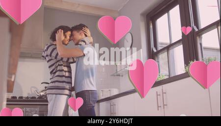 Bild von rosafarbenen Herzen über glücklichen, vielseitigen schwulen männlichen Paaren, die sich in der Küche umarmen Stockfoto
