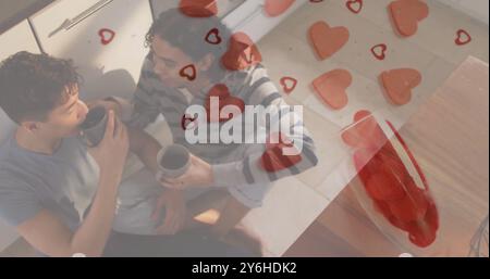 Bild von Rede Herzen über glückliche, vielfältige, schwule männliche Paare, die auf Küchenboden sitzen und Kaffee trinken Stockfoto