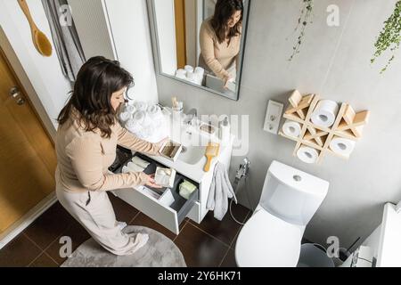 Glückliche junge Frau, die neben dem Frisiertisch steht und eine Schönheitskosmetik aus der Schublade herausnimmt. Stockfoto