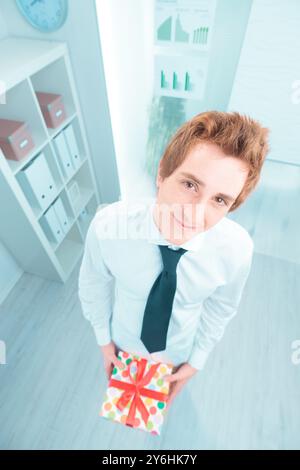 Junger Geschäftsmann in einem hellen Büro. Selbstbewusst ein Geschenk mit roter Schleife halten. Lächelnd und glücklich. Erfolg und Professionalität ausstrahlen. Feiert ein Stockfoto