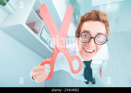 Ein lächelnder Geschäftsmann in einem Büro, der eine riesige rote Schere hält, strahlt Glück und Erfolg aus und schafft eine verspielte und humorvolle Unternehmensszene Stockfoto