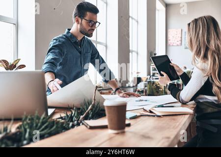Junge Geschäftsleute. Zwei junge, moderne Menschen in eleganter Freizeitkleidung, die Blueprint verwenden, während sie im kreativen Arbeitsbereich arbeiten Stockfoto