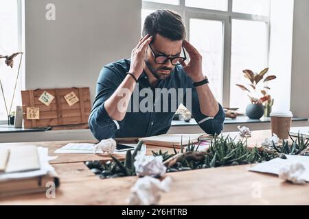 Schreckliche Kopfschmerzen. Müder junger Mann, der den Kopf in Händen hält, während er im Büro sitzt Stockfoto