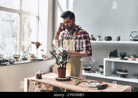 Ordnung annehmen. Hübscher junger Mann auf Schürze, der am Telefon spricht, während er in einem kleinen Gartencenter steht Stockfoto