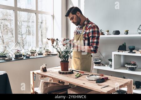 Alles wächst mit Liebe. Hübscher junger Mann auf Schürze, der mit Topfpflanzen arbeitet, während er in einem kleinen Gartenzentrum steht Stockfoto