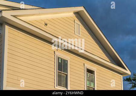 Sun malte die Seite eines amerikanischen Neubauhauses mit gelbem traditionellen Lap-Vinyl-Abstellgleis, gelbem Fensterrahmen und Giebelgiebel gelb Stockfoto