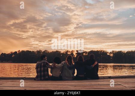 Leben voller Freundschaft. Rückansicht von Jugendlichen in Freizeitkleidung, die sich umarmen, während sie auf dem Pier sitzen Stockfoto