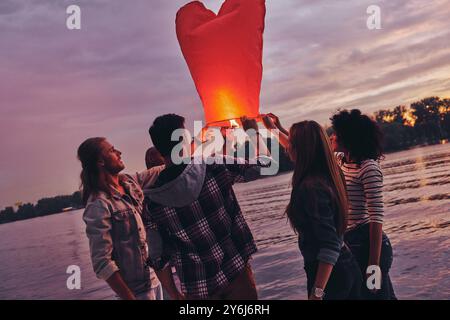 Leben voller Freundschaft. Gruppe junger Menschen in Freizeitkleidung, die die Himmelslaterne vorbereiten, während sie auf dem Pier stehen Stockfoto