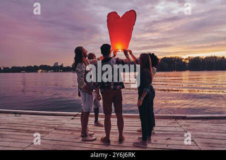 Spaß mit Freunden. Die ganze Länge der jungen Leute in Freizeitkleidung, die die Himmelslaterne vorbereiten, während sie auf dem Pier stehen Stockfoto