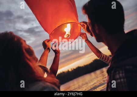 Glückliche Erinnerungen machen. Glückliche junge Menschen in Freizeitkleidung, die die Himmelslaterne halten, während sie in der Nähe des Flusses stehen Stockfoto