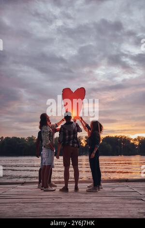 Wünschen Sie sich etwas! Die ganze Länge der jungen Leute in Freizeitkleidung, die die Himmelslaterne halten, während sie auf dem Pier stehen Stockfoto