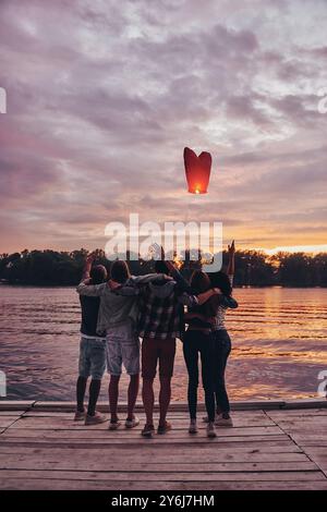 Fliegen Sie weg. Rückansicht der jungen Menschen in voller Länge, die sich umarmen und Gesten geben, während sie die schwimmende Himmelslaterne betrachten Stockfoto