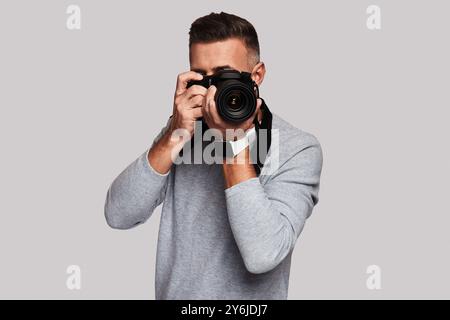 Schöne Aufnahme. Gut aussehender junger Mann, der dich fotografiert, während er vor grauem Hintergrund steht Stockfoto