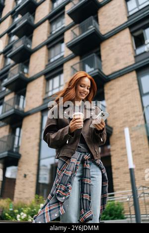 Vertikaler, flacher Blick auf die charmante Frau, die in der Stadtstraße steht, Kaffee hält und am Telefon scrollt, um Technologie und Mode mühelos zu kombinieren. Stockfoto