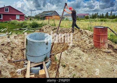 Der Baumeister platziert einen Betonring auf einen anderen und ebnet ihn mit einem Hebel mit einem Stahlrohr beim Bau einer Klärgrube. Stockfoto