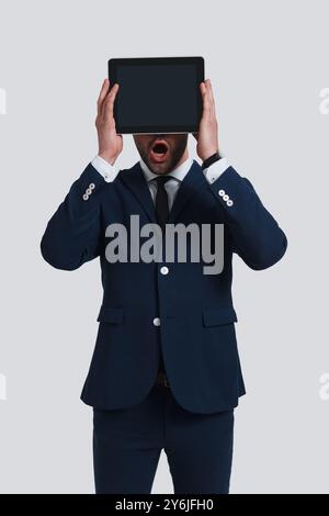 Echte Überraschung. Schockierter junger Mann im Vollanzug, der das Gesicht mit einem digitalen Tablet bedeckt und den Mund offen hält, während er vor grauem Hintergrund steht Stockfoto
