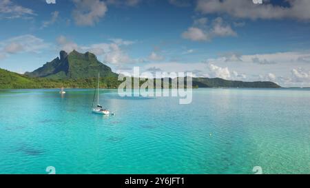 Zwei Segelboote Yachten ankern an der türkisfarbenen Küste der Bora Bora Lagune, grüne Berge ragen unter hellblauem Himmel. Abgelegenes wildes Naturparadies, exotische Sommerreise mit Luxus. Drohnenflug Stockfoto