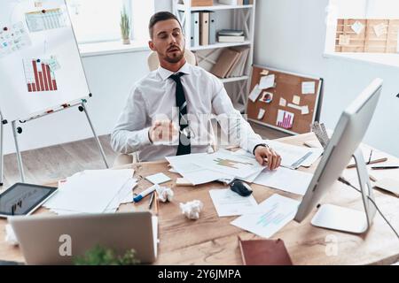 Im Büro stecken geblieben. Blick von oben auf einen müden jungen Mann in Formalkleidung, der mit Papieren arbeitet, während er im Büro sitzt Stockfoto