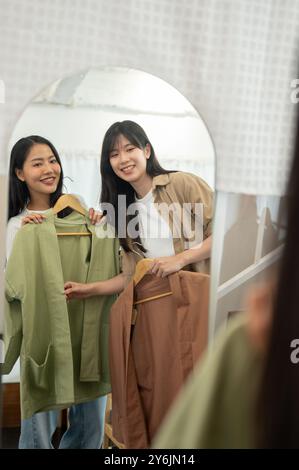 Zwei hübsche und fröhliche junge asiatische Freundinnen genießen es, gemeinsam in einem Geschäft einzukaufen, Kleidung anzuprobieren und sich im Spiegel zu bewundern. Stockfoto