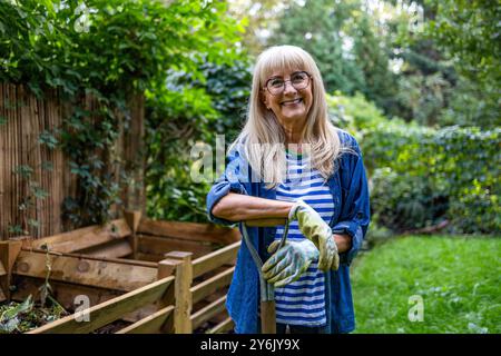 Eine ältere Frau, die mit einer Heugabel vor dem Komposthaufen im Garten steht Stockfoto