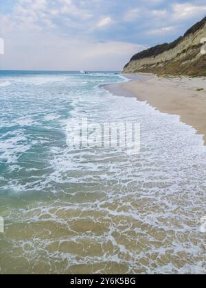 Eine ruhige Küstenszene mit türkisfarbenen Wellen, die sanft auf einen Sandstrand gleiten, umgeben von zerklüfteten Klippen. Perfekt für Natur, Reisen oder Meereslandschaft Stockfoto