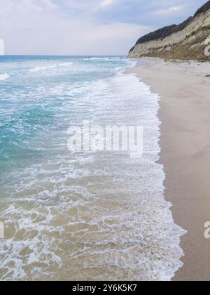 Eine ruhige Küstenszene mit türkisfarbenen Wellen, die sanft auf einen Sandstrand gleiten, umgeben von zerklüfteten Klippen. Perfekt für Natur, Reisen oder Meereslandschaft Stockfoto