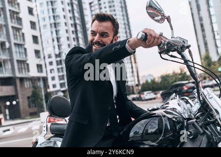 Lassen Sie uns eine Fahrt machen. Gut aussehender junger Mann im Vollanzug, der wegschaut und lächelt, während er draußen auf dem Motorrad sitzt Stockfoto