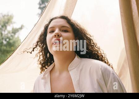 Eine selbstbewusste Frau in Übergröße steht inmitten des hohen Grases, getaucht in warmes Sonnenlicht und weiches Material. Stockfoto