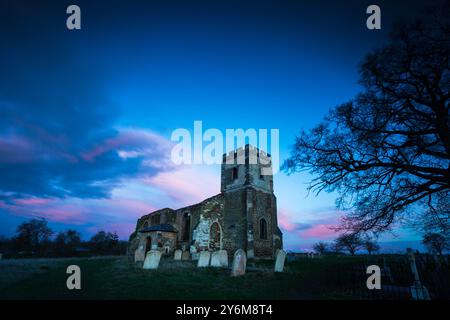 Eine alte Kirchenruine mit Grabsteinen unter einem lebendigen Himmel bei Sonnenuntergang, mit Silhouettenbäumen im Hintergrund. Stockfoto