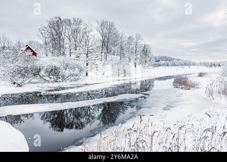 Der ruhige Fluss im Winter zeigt ein atemberaubendes Spiegelbild von schneebedeckten Bäumen und ein malerisches rotes Haus am Ufer, umgeben von dichter, frostiger Luft. Stockfoto