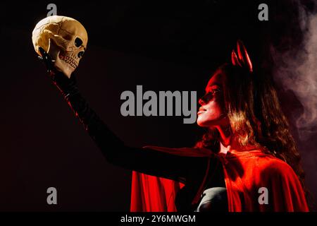 Eine umwerfende Frau mit rotem cape und Teufelshörnern hält einen Schädel und verkörpert Halloween-Geist. Stockfoto