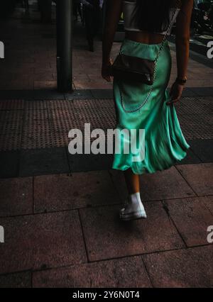 Vertikales Straßenfoto einer stilvollen jungen Frau, Mädchen in türkisfarbenem, seidenem Midirock aus Satin in Turnschuhen, die selbstbewusst auf dem Bürgersteig laufen. Street Fashion Stockfoto