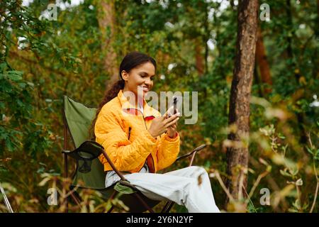 Eine junge Frau in leuchtenden Herbstfarben lächelt, als sie ihr Smartphone im Wald überprüft. Stockfoto