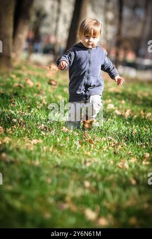 Ein kleines Kind läuft energisch durch einen lebendigen grünen Rasen mit gefallenen Blättern und genießt die Schönheit der Natur an einem sonnigen Tag im Park. Stockfoto