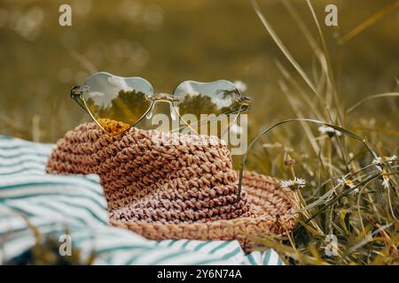 Strohhut im Boho-Stil und eine grüne Sonnenbrille in Herzform liegen auf einem grünen Rasen mit blühender Wiese. Sommerreisen. Sommer-Relax-Banner. Stockfoto