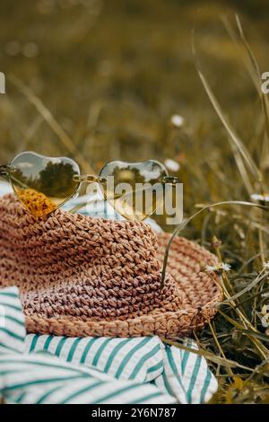 Strohhut im Boho-Stil und eine grüne Sonnenbrille in Herzform liegen auf einem grünen Rasen mit blühender Wiese. Sommerreisen. Sommer-Relax-Banner. Stockfoto