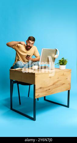 Junger emotionaler Mann, der mit Computer am Tisch sitzt und Online-Shopping über Handy vor blauem Studiohintergrund macht. Gute Angebote Stockfoto