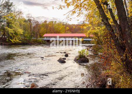 Traditionelle Holzbrücke mit rotem Dach, die einen Fluss mit bewaldeten Ufern überspannt und die Herbstfarben bei Sonnenuntergang überragt Stockfoto