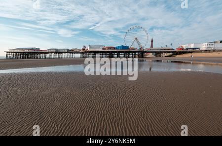 Ruhige Strandszene mit Pier, Riesenrad und Reflexionen auf nassem Sand unter klarem Himmel. Stockfoto