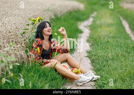 Eine junge Frau, die am Rande eines Feldweges zwischen grünem Gras und reifem Weizenfeld sitzt. Sie lacht fröhlich und hält einen Haufen Trauben in sich Stockfoto