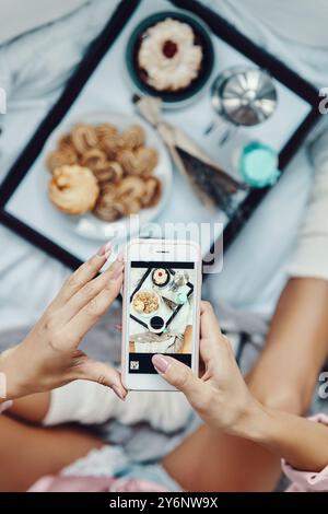 Nahaufnahme einer jungen Frau im Pyjama, die ein flaches Frühstück fotografiert, während sie zu Hause im Bett ruht Stockfoto