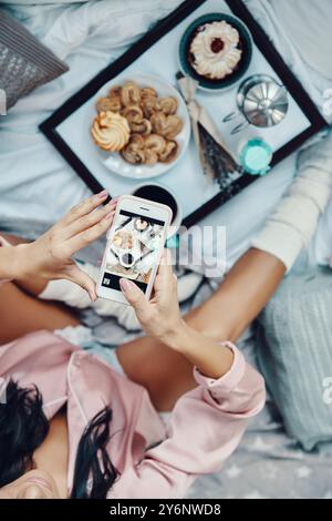 Nahaufnahme einer jungen Frau im Pyjama, die ein flaches Frühstück fotografiert, während sie zu Hause im Bett ruht Stockfoto