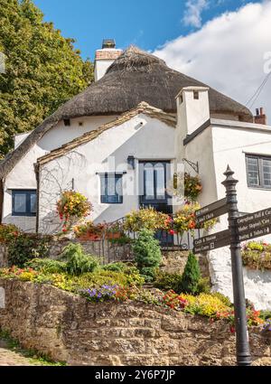 Ein Haus mit einem Strohdach steht auf einem Eckgrundstück mit einem Felsbrocken voller farbenfroher Blumen. Ein Wegweiser ist im Vordergrund und ein Himmel mit Wolke i Stockfoto