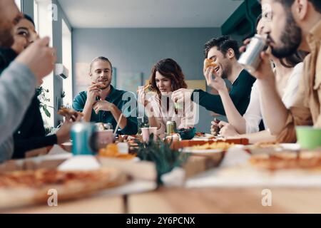Pizza mit Freunden genießen. Gruppe junger Leute in Freizeitkleidung, die Pizza essen und lächeln, während sie eine Dinnerparty im Haus feiern Stockfoto