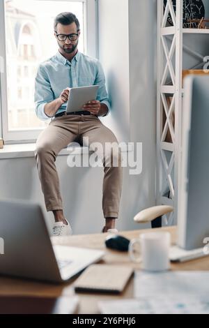 Immer verfügbar. Junger, moderner Geschäftsmann, der mit einem digitalen Tablet arbeitet, während er im Büro auf der Fensterbank sitzt Stockfoto