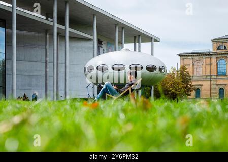 Ein junger Mann sitzt in einem Liegestuhl auf dem Rasen vor der Pinakothek der Moderne und der Kunstinstallation Futuro House von Matti Suuronen, München Stockfoto