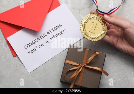 Frau mit goldener Medaille, Karte mit Phrase Herzlichen Glückwunsch Sie sind Gewinner, Geschenkbox und Umschlag auf grauem Marmortisch, Blick von oben Stockfoto